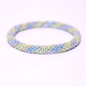 Puede incluir: Una pulsera de cuentas en tonos azul claro, verde y dorado. La pulsera está hecha de pequeñas cuentas espaciadas uniformemente, creando un patrón en espiral. La pulsera es circular y parece hecha a mano.