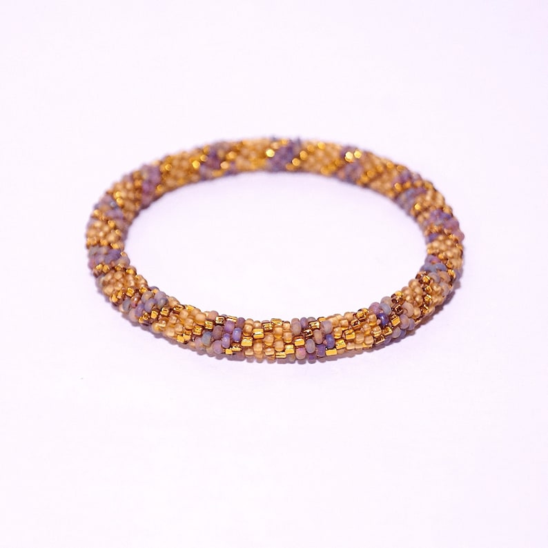 Op de afbeelding: Een close-up van een kralenarmband met een gouden en paarse kleurstelling. De armband is gemaakt van kleine, ronde kralen die in een spiraalvorm met elkaar zijn verweven.