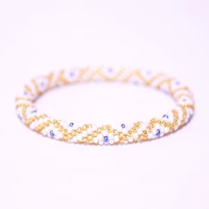 Puede incluir: Pulsera de cuentas blancas y doradas con detalles azules. La pulsera está hecha de cuentas pequeñas y tiene un patrón de chevron.