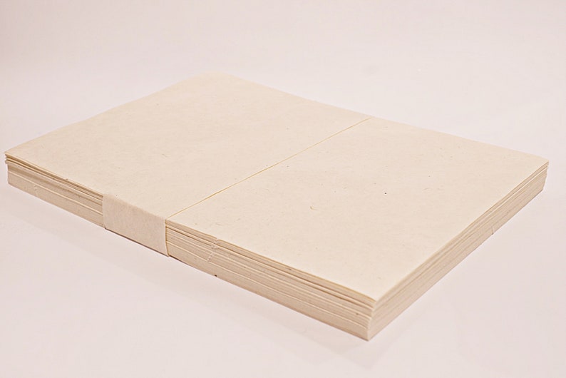 K&ouml;nnte beinhalten: Ein Stapel cremefarbener Papierbl&auml;tter, zusammengebunden mit einem passenden Papierband. Das Papier hat eine glatte Textur und ist ordentlich gestapelt, was auf ein Schreibwaren- oder Kunstbedarfsprodukt hindeutet.