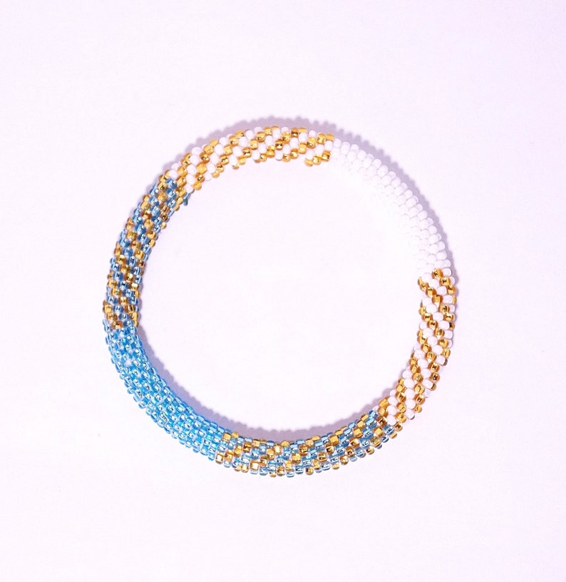 May include: A beaded bracelet with a spiral design. The bracelet features a gradient of colors, transitioning from light blue to gold and white. The beads are small and tightly woven, creating a textured surface. The bracelet is circular and appears to be handmade.