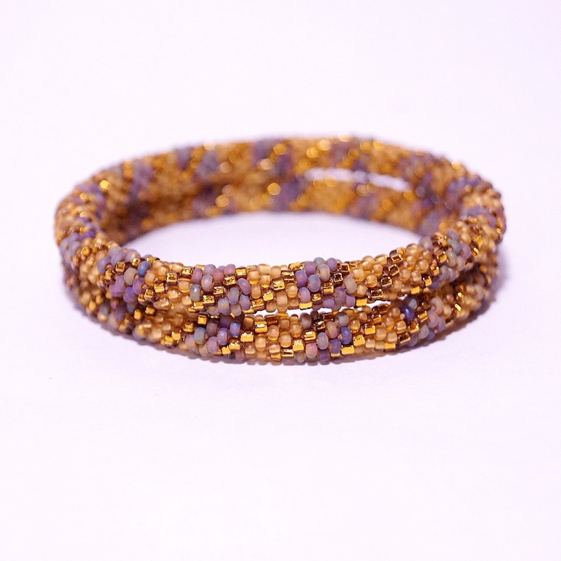 Op de afbeelding: Een dubbele rij kralen armband met een gouden en paarse kleurstelling. De armband is gemaakt van kleine, ronde kralen die in een spiraalvorm met elkaar zijn verweven.