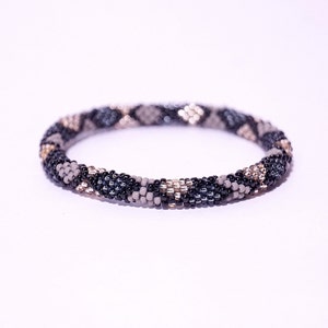Puede incluir: Un brazalete de cuentas de estilo enrollado, con un patrón de cuentas negras, grises y doradas. El brazalete es circular y parece hecho a mano, con una superficie texturizada y detallada. Las cuentas son pequeñas y están tejidas apretadamente. El diámetro es de unos 6 cm.