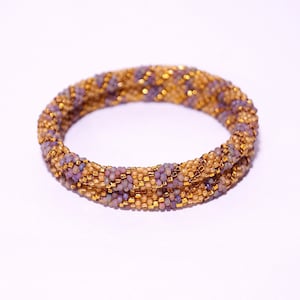 Op de afbeelding: Een kralenarmband met een paarse en gouden kleurstelling. De armband is gemaakt van kleine, ronde kralen die in een spiraalvorm met elkaar zijn verweven.