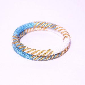 May include: A beaded bracelet with a spiral design. The bracelet features alternating sections of light blue, gold, and white beads. The beads are tightly strung together, creating a textured surface. The bracelet is shown on a white background.