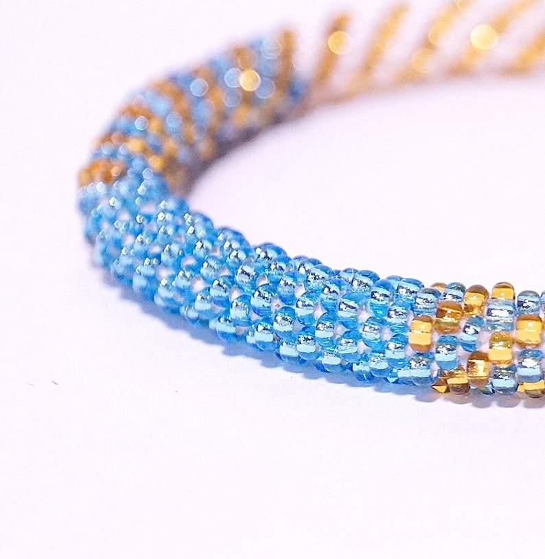 May include: Close-up of a beaded bracelet. The bracelet features a spiral design with alternating sections of light blue and gold beads. The beads are small and round, creating a textured surface. The background is white.