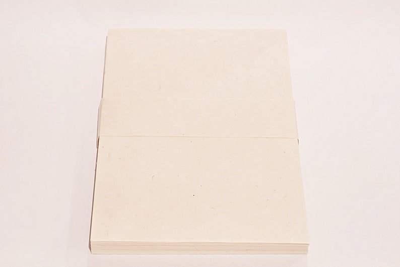 K&ouml;nnte beinhalten: Ein Stapel cremefarbenes Papier, wahrscheinlich Briefpapier oder Notizkarten, wird vor einem wei&szlig;en Hintergrund gezeigt. Das Papier scheint aus einem strukturierten, hochwertigen Material mit sichtbarer Maserung zu bestehen. Der Stapel ist ordentlich angeordnet.
