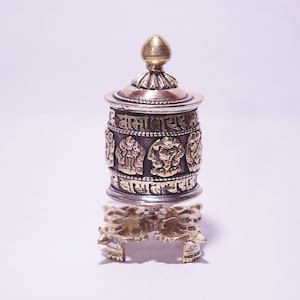 May include: A silver and gold prayer wheel with intricate carvings and script. The cylindrical body features detailed relief images and is topped with a gold finial. It sits on a decorative gold stand.