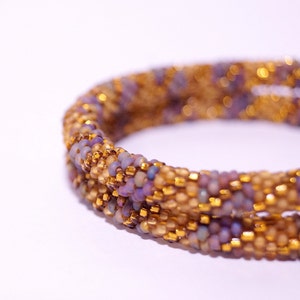 Op de afbeelding: Een close-up van een kralenarmband met een spiraalvormig ontwerp. De armband is gemaakt met kleine, iriserende paarse en gouden kralen.