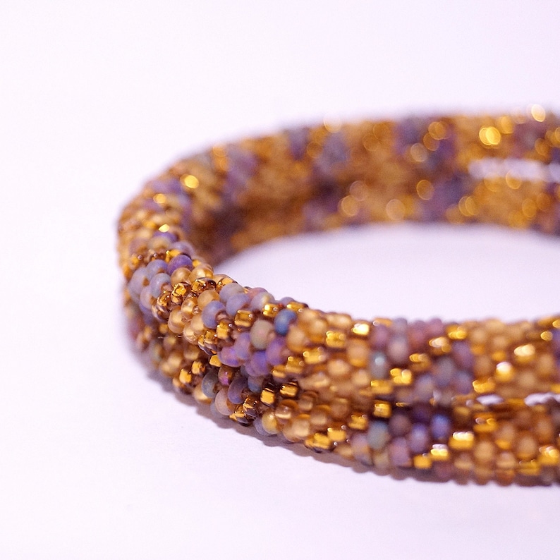 Op de afbeelding: Een close-up van een kralenarmband met een spiraalvormig ontwerp. De armband is gemaakt met kleine, iriserende paarse en gouden kralen.