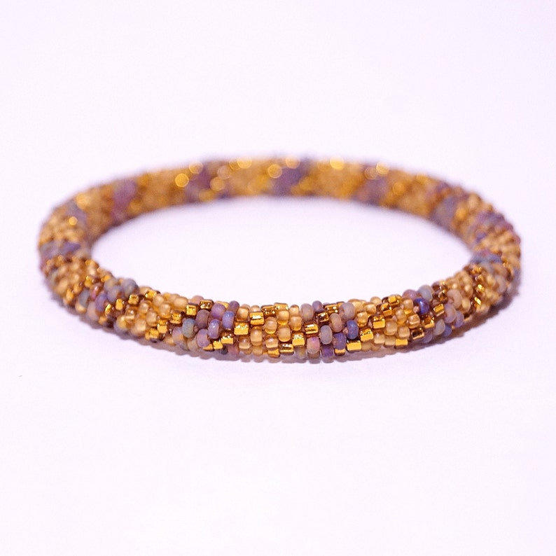 Op de afbeelding: Een kralen armband met een gouden en paarse kleurstelling. De armband is gemaakt van kleine, ronde kralen die in een spiraalvorm met elkaar zijn verweven.