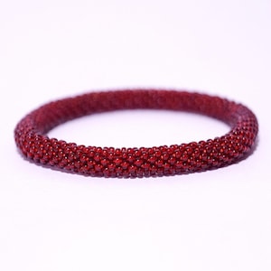 Op de afbeelding: Een bordeauxrode kralenarmband. De armband is gemaakt van kleine, ronde kralen, die strak aan elkaar zijn geregen om een ​​stevige band te vormen. De kralen hebben een glanzende afwerking, die het licht reflecteert. De armband heeft een dieprode kleur en staat tegen een witte achtergrond.