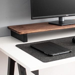 Pode incluir: Um suporte para monitor de madeira marrom escuro com suportes de metal preto, um teclado preto, um tablet preto e um tapete de rato preto em uma mesa branca.