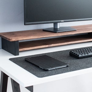 May include: A white desk with a black metal frame and a dark grey mouse mat. A black keyboard and a black tablet are on the mouse mat. A wooden monitor stand with a black metal frame sits on the desk. The monitor stand has a shelf underneath. A black computer monitor with the Dell logo is on the stand.