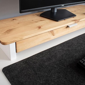 Pode incluir: Um suporte de monitor de madeira com acabamento em madeira natural e base branca. O suporte tem uma gaveta com puxador prateado. Um monitor de computador preto, um rato branco e um teclado preto estão na secretária.