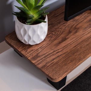 May include: A wooden TV stand with black legs and a dark brown stain. A white planter with a green succulent plant sits on the top of the stand.