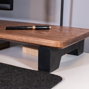 May include: A wooden monitor stand with black legs. The stand is made of dark wood and has a smooth, polished finish. The stand is raised off the surface by four black legs.