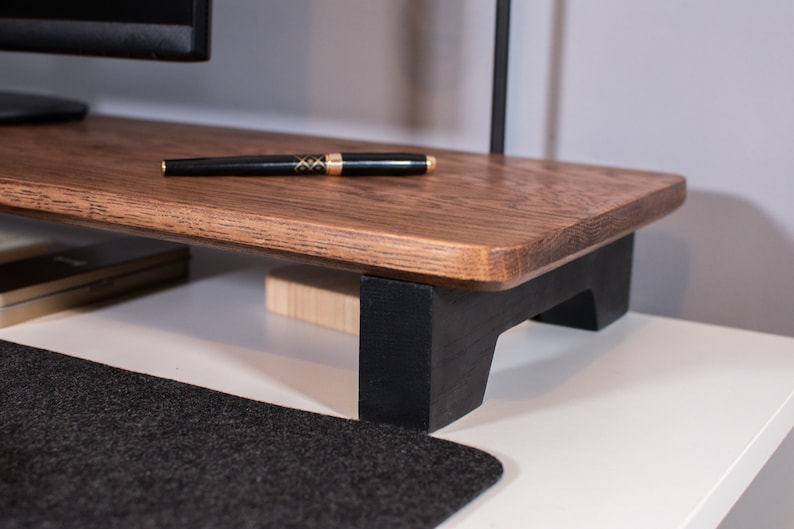 May include: A wooden monitor stand with black legs. The stand is made of dark wood and has a smooth, polished finish. The stand is raised off the surface by four black legs.