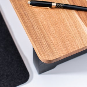 Pu&ograve; includere: Un supporto per laptop in legno con base nera e piano in legno naturale. Una penna nera &egrave; appoggiata sulla parte superiore del supporto.