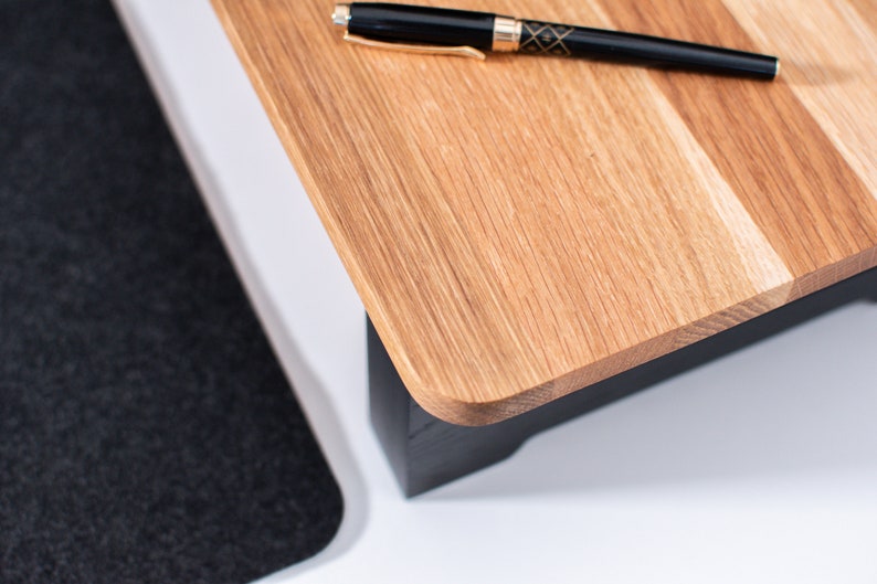 Pu&ograve; includere: Un supporto per laptop in legno con base nera e piano in legno naturale. Una penna nera &egrave; appoggiata sulla parte superiore del supporto.