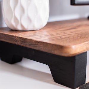 May include: A wooden monitor stand with black painted legs. The stand is made of natural wood with a light brown finish. The stand is raised off the surface with a gap between the bottom of the stand and the surface.