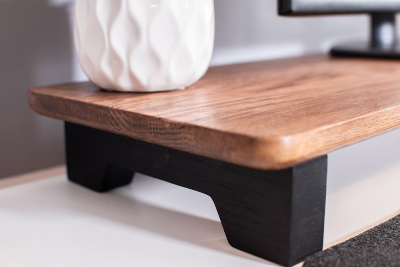 May include: A wooden monitor stand with black painted legs. The stand is made of natural wood with a light brown finish. The stand is raised off the surface with a gap between the bottom of the stand and the surface.