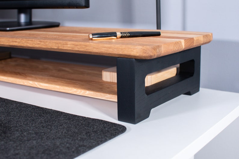 Pu&ograve; includere: Un supporto per monitor in legno con una base verniciata di nero e uno scaffale. Il supporto &egrave; in legno chiaro e ha una finitura naturale. Una penna &egrave; appoggiata sullo scaffale superiore.