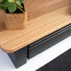 K&ouml;nnte beinhalten: Ein TV-St&auml;nder aus Holz mit einer schwarzen Schublade und einem schwarzen Sockel. Die Oberseite des St&auml;nders ist aus hellem Holz und hat eine nat&uuml;rliche Oberfl&auml;che. Eine kleine Topfpflanze steht auf der Oberseite des St&auml;nders.