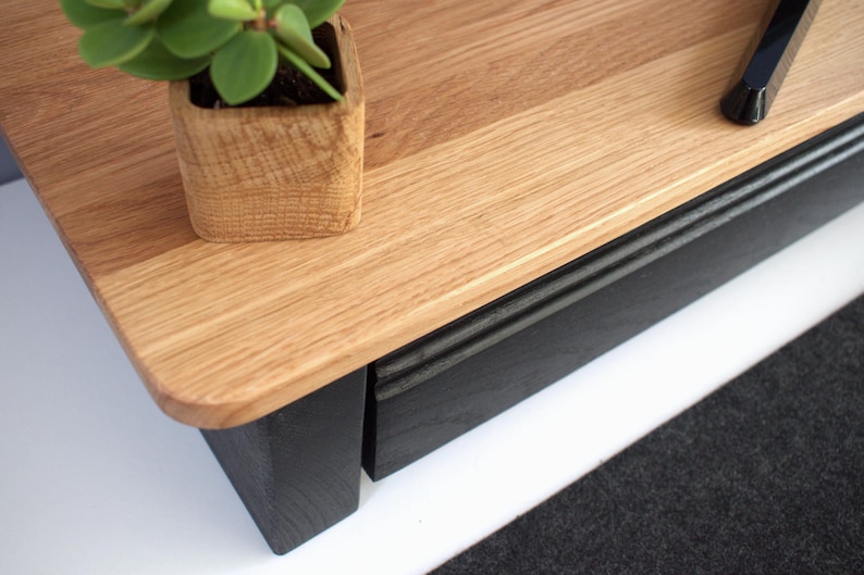 K&ouml;nnte beinhalten: Ein TV-St&auml;nder aus Holz mit einer schwarzen Schublade und einem schwarzen Sockel. Die Oberseite des St&auml;nders ist aus hellem Holz und hat eine nat&uuml;rliche Oberfl&auml;che. Eine kleine Topfpflanze steht auf der Oberseite des St&auml;nders.