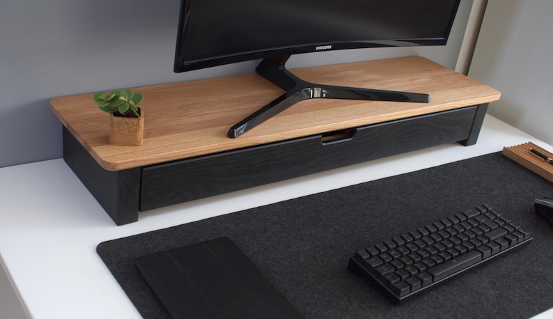 K&ouml;nnte beinhalten: Ein Monitorst&auml;nder aus Holz mit einer schwarzen Schublade, einer kleinen Topfpflanze und einer schwarzen Tastatur auf einer schwarzen Schreibtischmatte.