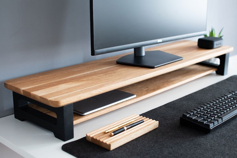 Pu&ograve; includere: Un supporto per monitor in legno con una cornice in metallo nero e uno scaffale inferiore. Un laptop &egrave; sullo scaffale e un portapenne &egrave; sulla scrivania sotto il supporto.
