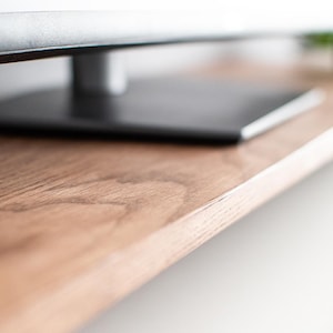 May include: Close-up of a wooden TV stand with a dark brown finish. The stand has a smooth, natural wood grain pattern. The top shelf is empty, while the bottom shelf is out of focus.