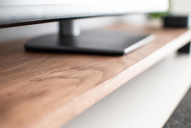 May include: Close-up of a wooden TV stand with a dark brown finish. The stand has a smooth, natural wood grain pattern. The top shelf is empty, while the bottom shelf is out of focus.