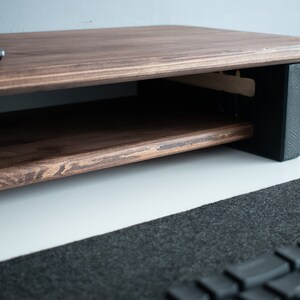 May include: A dark brown wooden monitor stand with a shelf underneath. The stand has black supports on the sides and is sitting on a white surface.