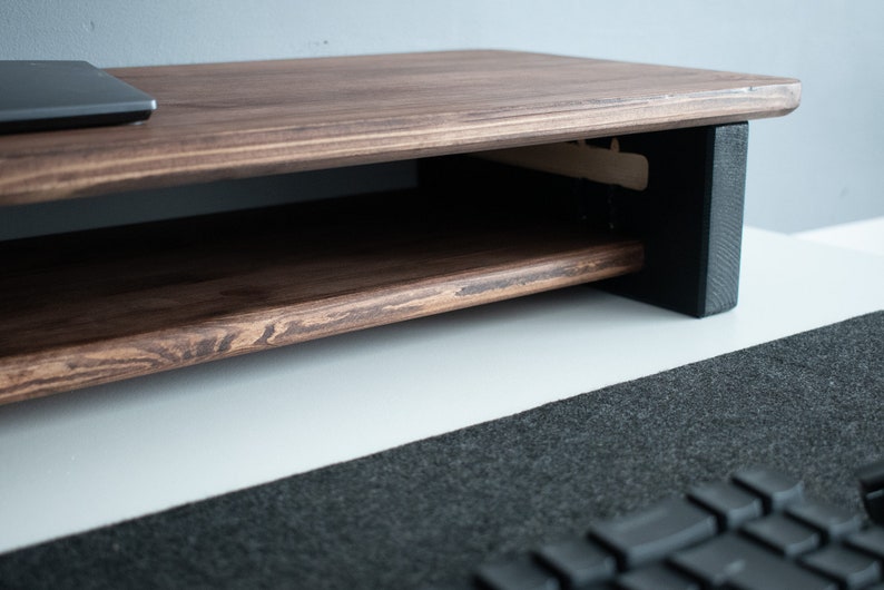 May include: A dark brown wooden monitor stand with a shelf underneath. The stand has black supports on the sides and is sitting on a white surface.