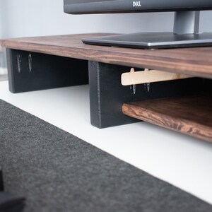 May include: A wooden monitor stand with a dark brown finish and a lower shelf. The stand has black supports and is sitting on a white desk with a gray mouse pad.