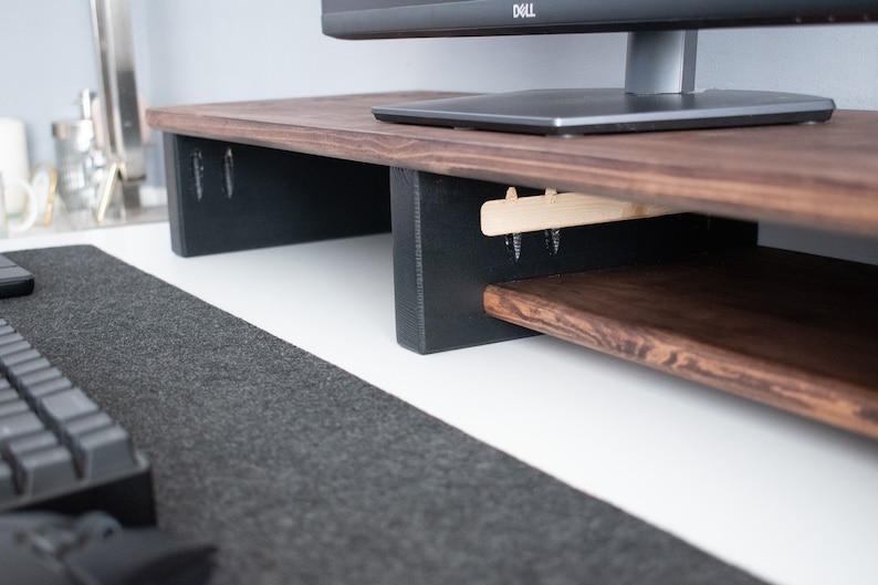 May include: A wooden monitor stand with a dark brown finish and a lower shelf. The stand has black supports and is sitting on a white desk with a gray mouse pad.