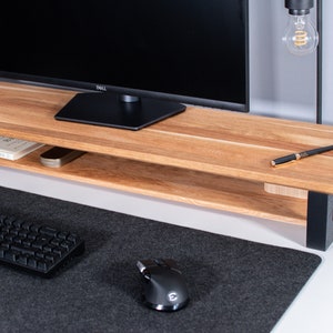 Pu&ograve; includere: Un supporto per monitor in legno con una cornice in metallo nero e un ripiano inferiore. Il supporto &egrave; su una scrivania bianca con un tappetino per mouse nero, una tastiera nera e un mouse nero.