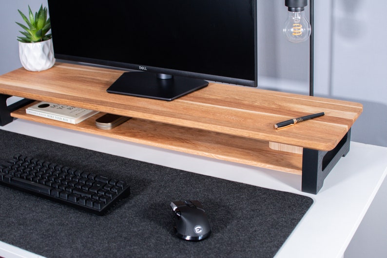 Pu&ograve; includere: Un supporto per monitor in legno con una cornice in metallo nero e un ripiano inferiore. Il supporto &egrave; su una scrivania bianca con un tappetino per mouse nero, una tastiera nera e un mouse nero.