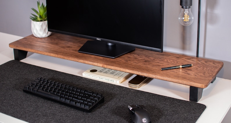 May include: A wooden monitor stand with black metal legs, a black keyboard, a black mouse, and a book titled "Make Your Bed" on the shelf below the stand.