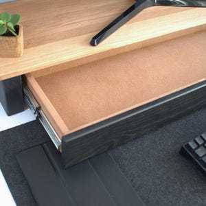 K&ouml;nnte beinhalten: Ein schwarzer Holz-Schreibtisch-Aufsatz mit einer Schublade, die offen ist und ein hellbraunes Inneres zeigt. Der Aufsatz steht auf einer schwarzen Filzunterlage. Eine schwarze Tastatur liegt auf der Unterlage.