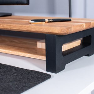 Pu&ograve; includere: Un supporto per monitor in legno con una base verniciata di nero e uno scaffale. Il supporto &egrave; in legno naturale con una finitura marrone chiaro. Una penna &egrave; appoggiata sullo scaffale superiore.