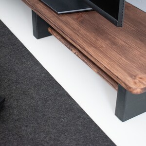 May include: A dark brown wooden monitor stand with black painted legs. The stand is on a white surface and a black keyboard and mouse are on a dark gray surface to the left.