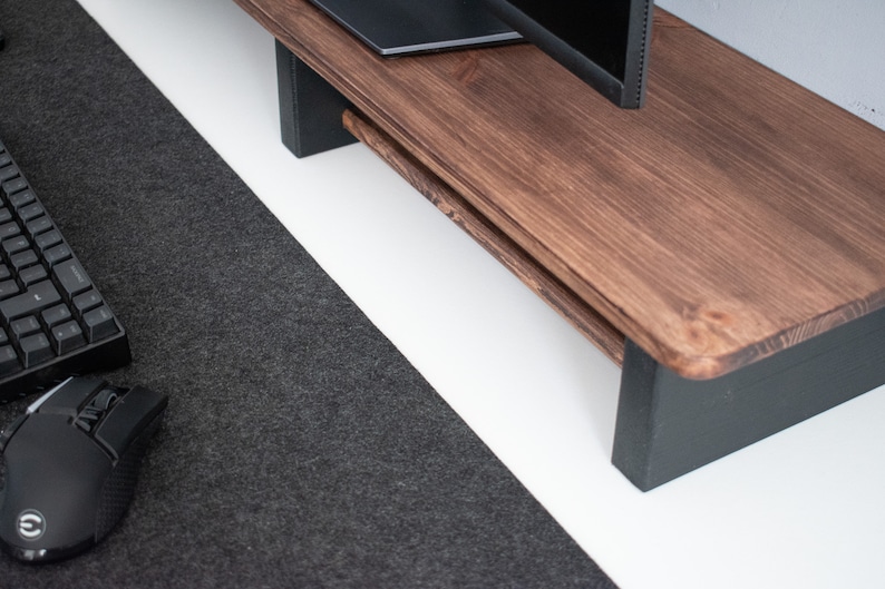 May include: A dark brown wooden monitor stand with black painted legs. The stand is on a white surface and a black keyboard and mouse are on a dark gray surface to the left.