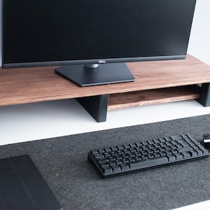 May include: A wooden monitor stand with a black monitor on top. The stand has a shelf underneath for storage. A black keyboard and mouse are on a gray desk mat.