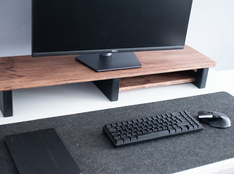 May include: A wooden monitor stand with a black monitor on top. The stand has a shelf underneath for storage. A black keyboard and mouse are on a gray desk mat.