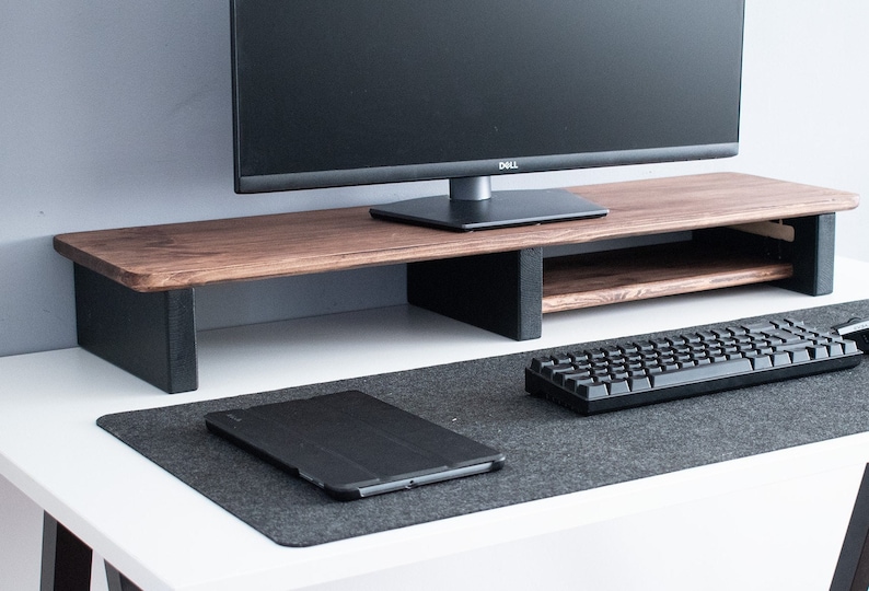 May include: A wooden monitor stand with a black shelf underneath. The stand is on a white desk with a black keyboard and a black tablet on a gray desk mat.
