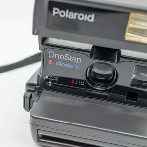Peut inclure: Un appareil photo Polaroid OneStep Close Up gris avec une sangle noire. L'appareil photo est doté d'un flash intégré et d'un objectif macro.