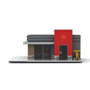 Peut inclure: Une maquette de bâtiment en blocs de construction, avec une façade rouge ornée d'un smiley jaune et d'une porte noire. Le bâtiment a un toit gris et un côté marron clair et gris. Un vélo jaune est garé à côté.
