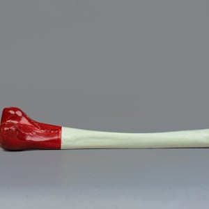 May include: A red and white painted bone model. The bone is curved and has a rounded end. The red paint covers the rounded end and part of the bone. The rest of the bone is painted white.